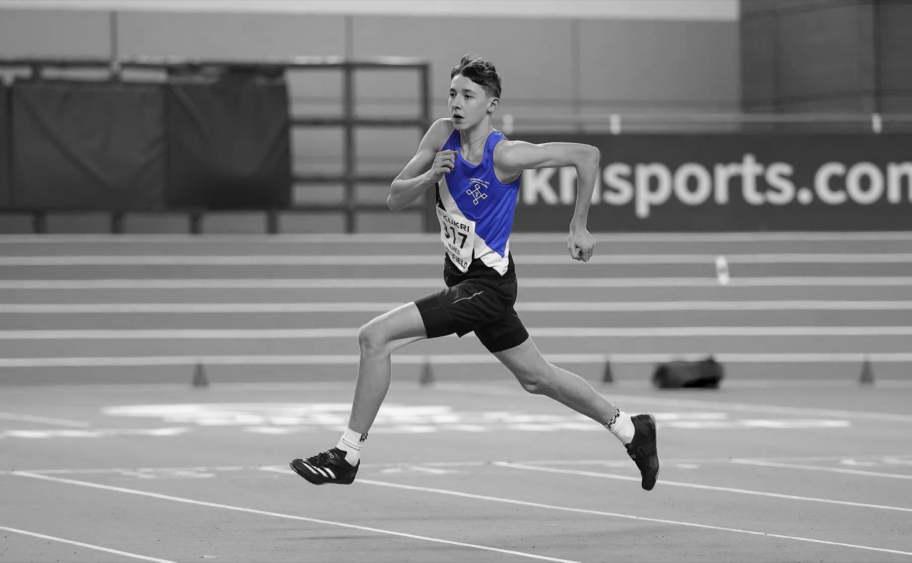 A Kingston & Poly athlete in the blue and black vest sprinting on an indoor track